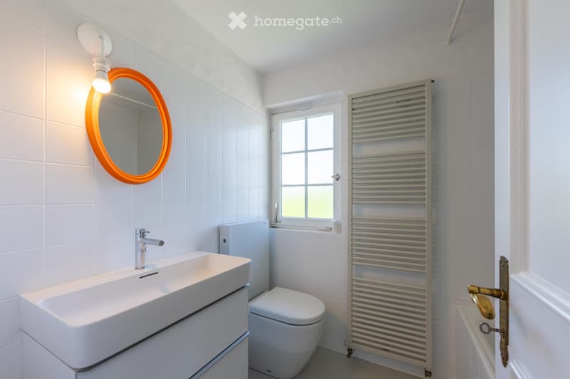 Modern bathroom with white tiles, a large sink with a chrome faucet, a round mirror with an orange frame, a radiator, and a toilet.