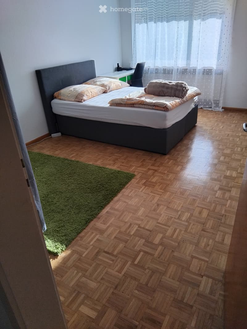 Bedroom with wooden herringbone flooring, a large bed with pillows and blankets, a green rug, a desk with a chair, and a window with sheer curtains.