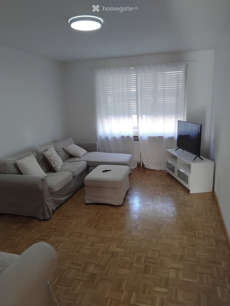 Living room with wooden flooring, a grey sectional sofa, an ottoman, a TV stand with a flat-screen TV, and a remote control on the ottoman.