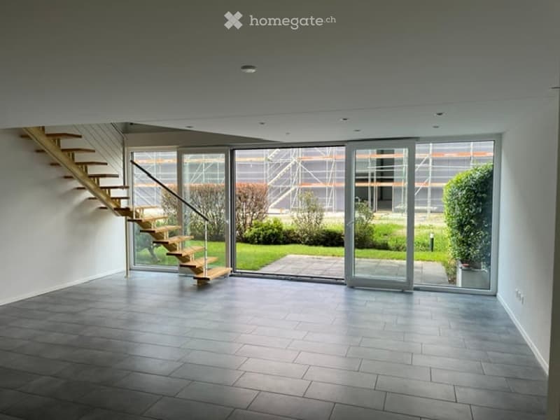 Open-plan space with a wooden staircase leading to an upper level, large glass doors opening to a garden, and a tiled floor.