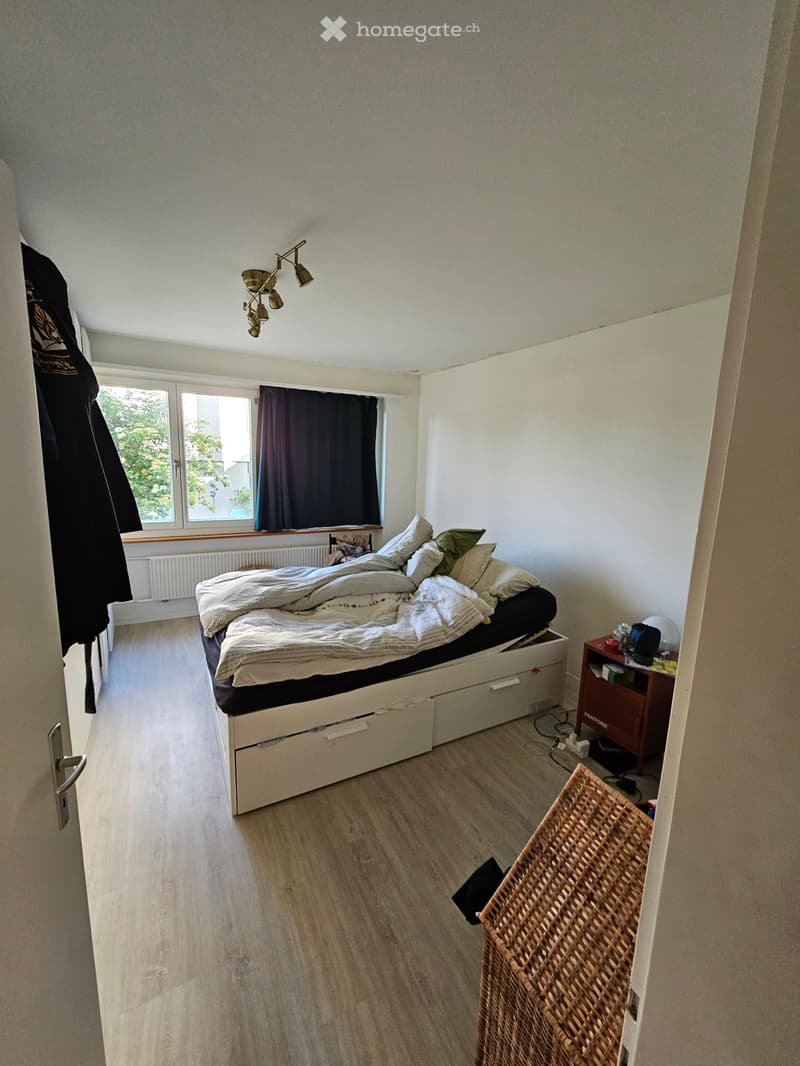 Cozy bedroom with a white bed, nightstand, wicker basket, and a lamp. The room features wooden flooring and large windows with dark curtains.