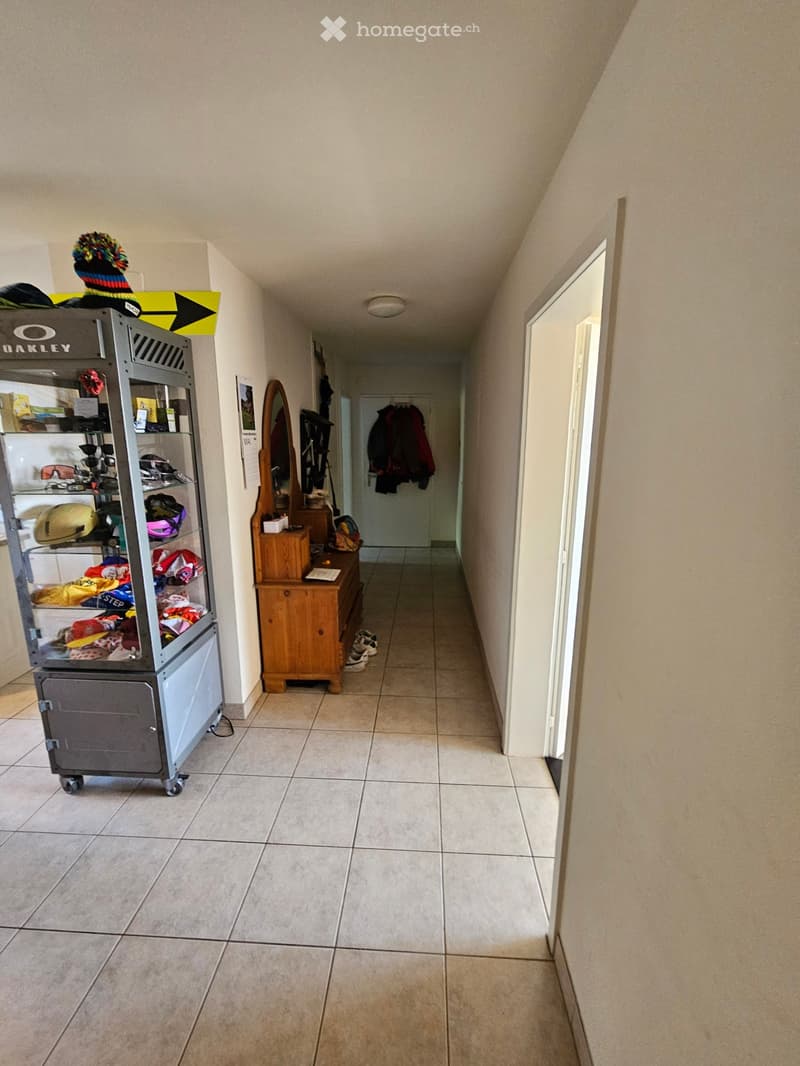 A narrow hallway with tiled flooring, beige walls, and a wooden dresser with a mirror. A metal cart with shelves is visible on the left side, and a coat rack with jackets is seen at the end of the hallway.