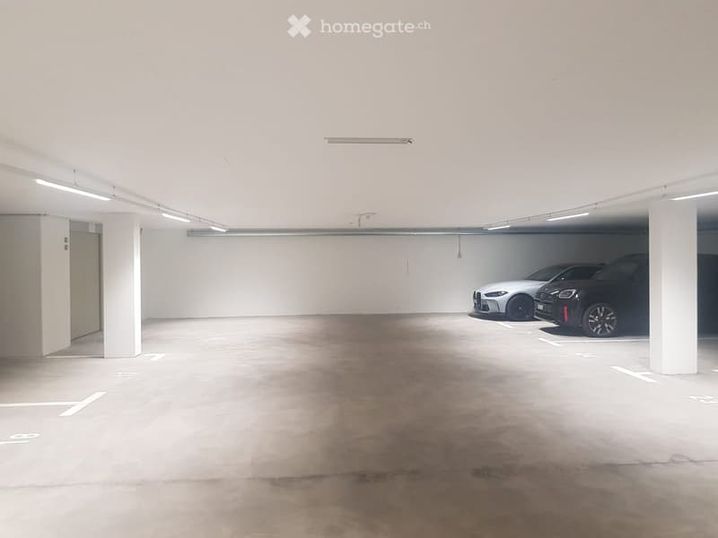 Underground parking area with two parked cars, concrete floor, white pillars, and fluorescent lighting.