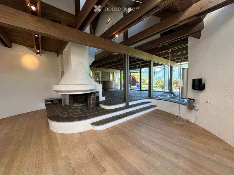 Modern living room with exposed wooden beams, a large fireplace, hardwood flooring, and large windows offering a view of the outdoor area.