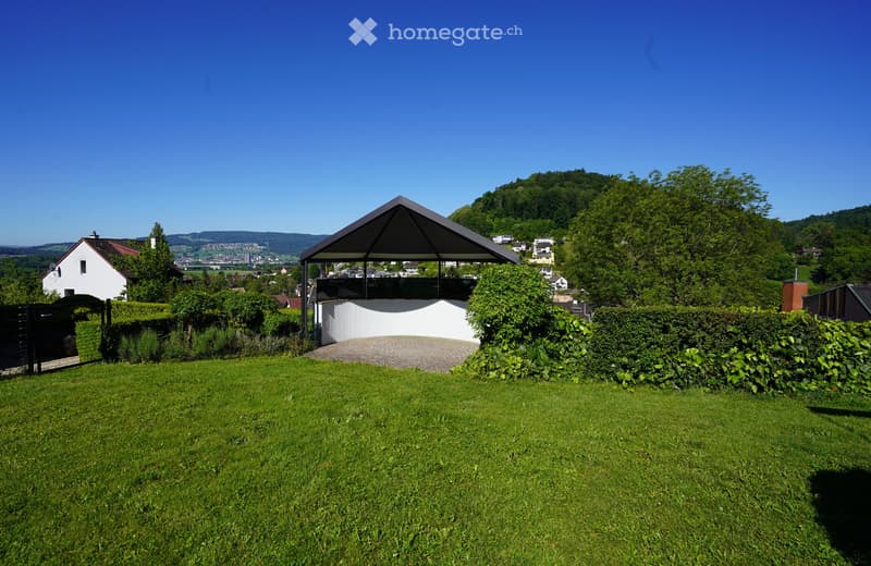 A well-maintained garden with a paved driveway leading to a modern, angular carport. The area is surrounded by lush green hedges and trees, with a scenic view of a town and hills in the background.
