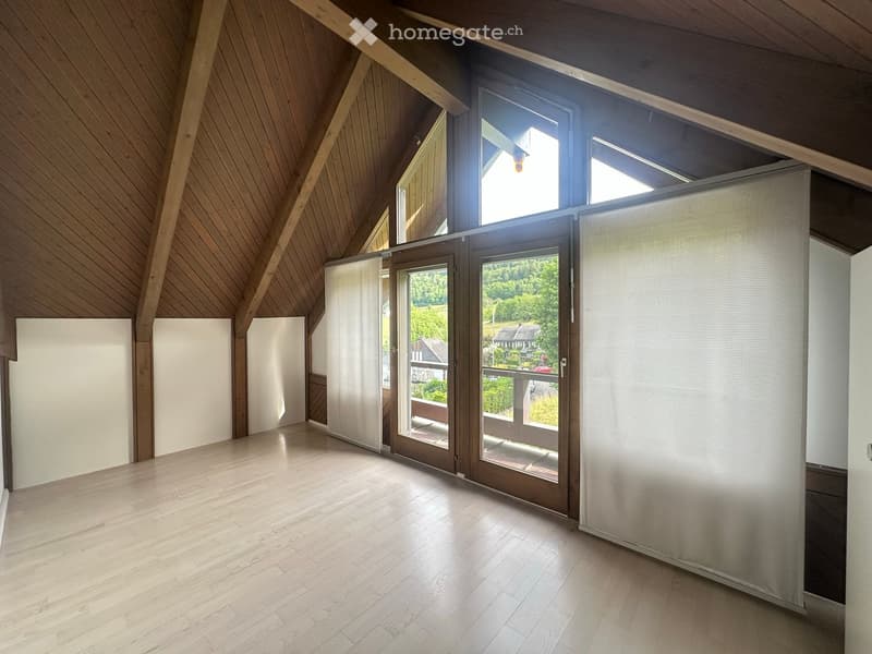 Bright, spacious attic room with wooden walls and floor, large windows and sliding doors leading to a balcony, white paneling on one wall.