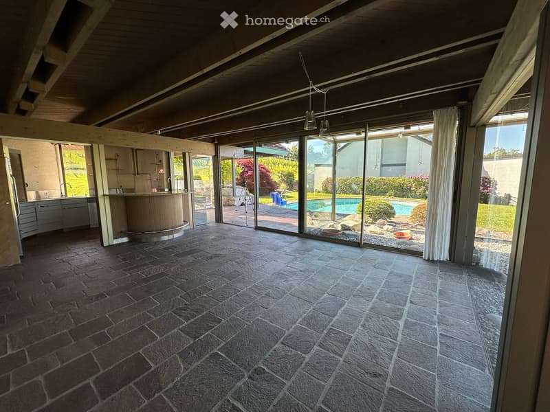 Open-plan space with stone flooring, large glass sliding doors leading to a pool area, visible kitchen area with cabinets and a curved bar counter.