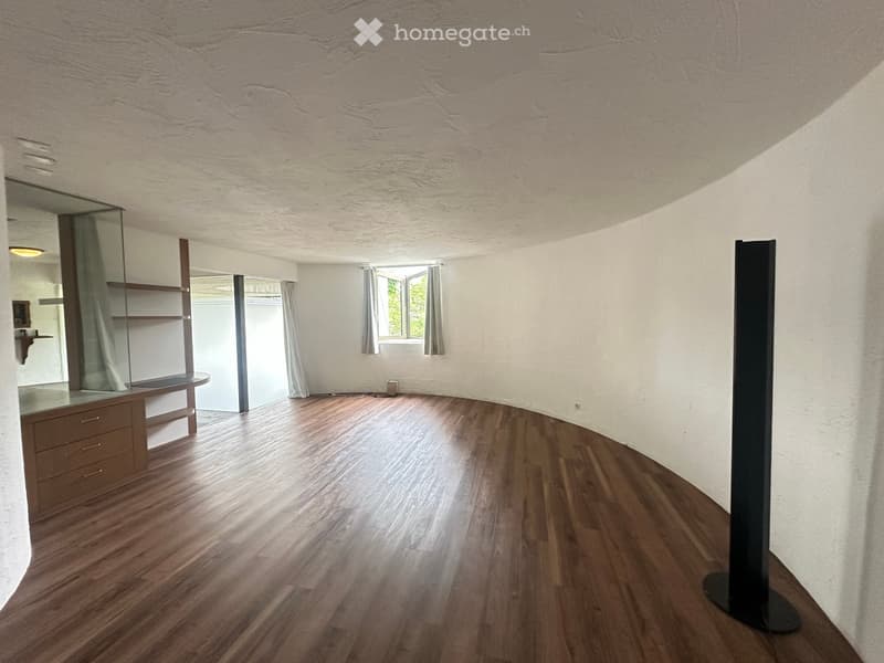 Empty room with wooden flooring, white textured walls, and a large window with curtains. There is a glass cabinet with shelves and a small table visible in the background.