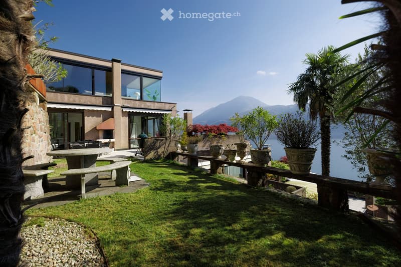A modern house with a spacious outdoor garden area, featuring a stone table, benches, potted plants, and a scenic view of a lake and mountains.