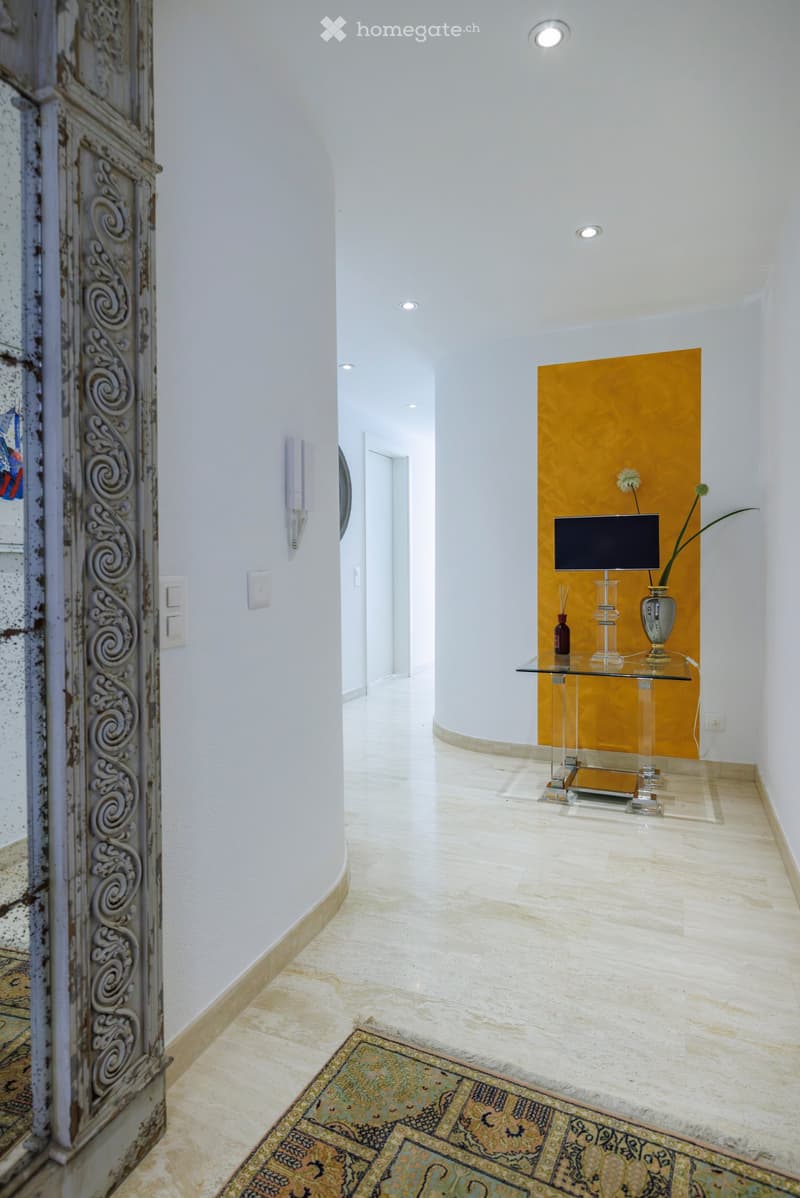 A modern hallway with white walls, a glass table, a vase with flowers, and a screen on a yellow accent wall.