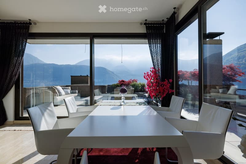 Modern dining area with white table and chairs, sofa, and vase, large windows offering scenic mountain view.