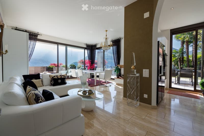 Modern living room with large windows offering a scenic view, white leather sofa, glass coffee table, chandelier, dining area with white table and chairs, decorative vases, and curtains.