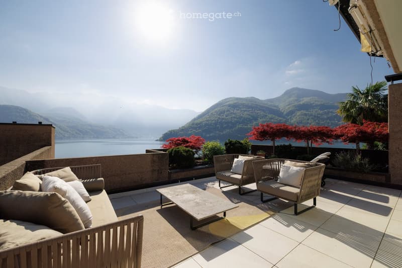 A spacious balcony with a scenic lake and mountain view, featuring wicker sofas, a coffee table, and decorative pillows.