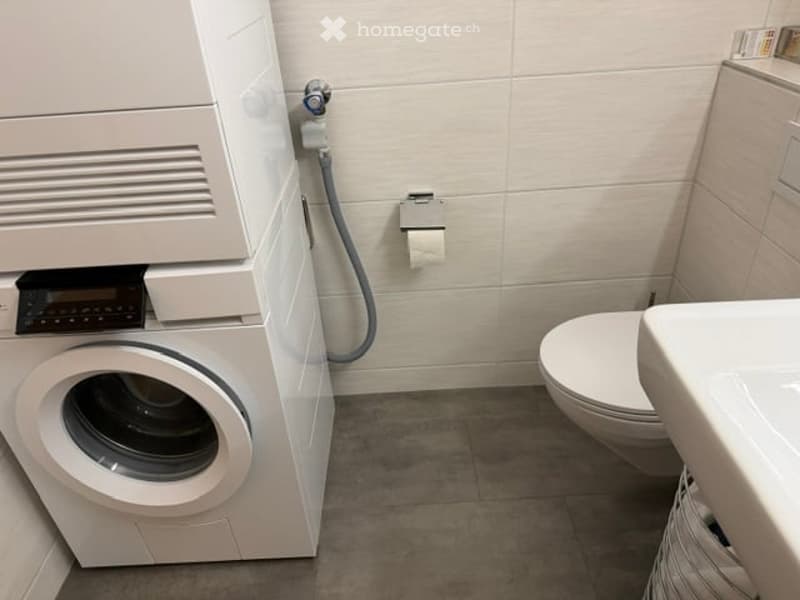 Modern bathroom with a toilet, sink, and washing machine. Light-colored tiles and a clean, minimalist design.