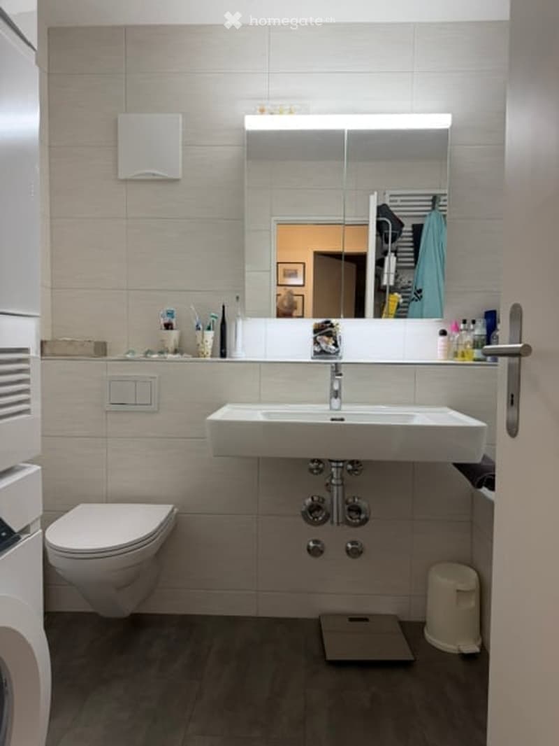 Modern bathroom with a white sink, large mirror, toilet, and various toiletries on the counter. The walls are tiled in a light beige color, and the floor is dark grey.