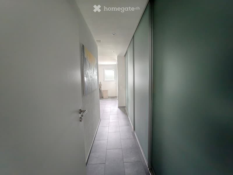 A clean, modern hallway with tiled flooring, white walls, and recessed lighting. A door is visible on the left, and a small room with a window is visible at the end of the corridor.