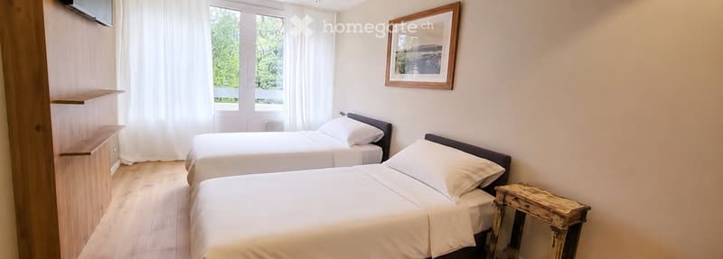 Cozy bedroom with two single beds, white linens, wooden nightstand, and a framed picture on the wall. Large windows with white curtains offer a view of greenery outside.