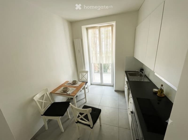 A compact kitchen and dining area with white walls, tiled flooring, and a black countertop. A small wooden dining table with four white chairs is set near a sliding glass door, and a sink with a faucet is visible on the right side.