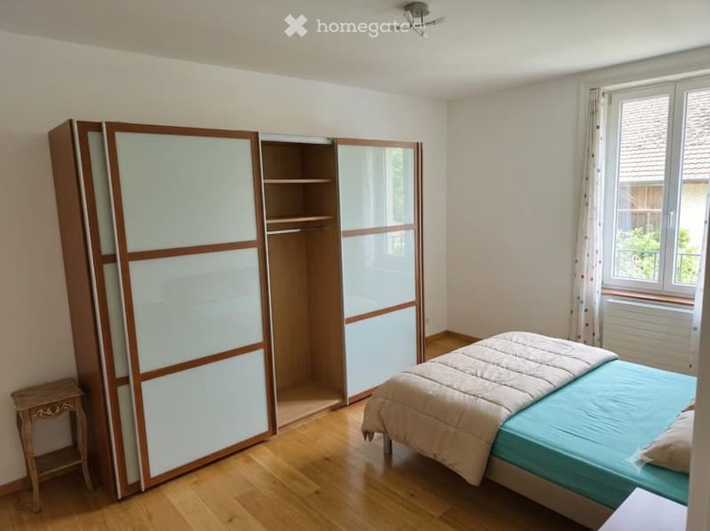 Bedroom with wooden floor, large window, bed with blue sheets, wardrobe with mirrored doors, and a small nightstand.
