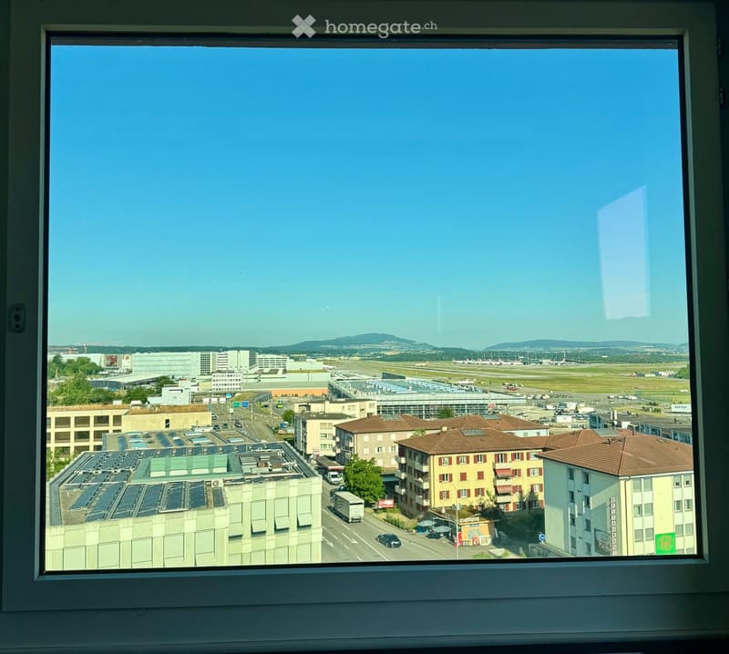 View from a window showing an airport with runways, buildings, and distant mountains under a clear blue sky.