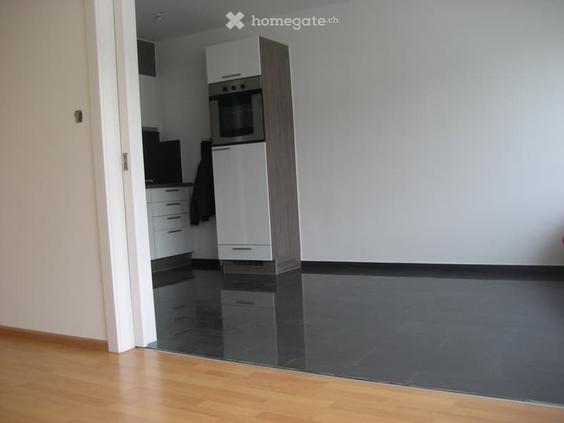 Empty room with wooden flooring and a doorway leading to a kitchen area with white cabinets, a built-in oven, and a countertop.