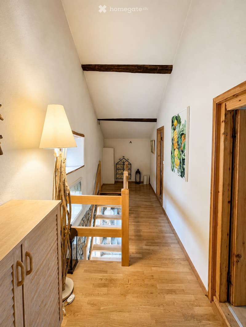 A well-lit hallway with wooden flooring, white walls, and a slanted ceiling. Features include a wooden railing, a tall floor lamp, and several framed artworks on the walls.