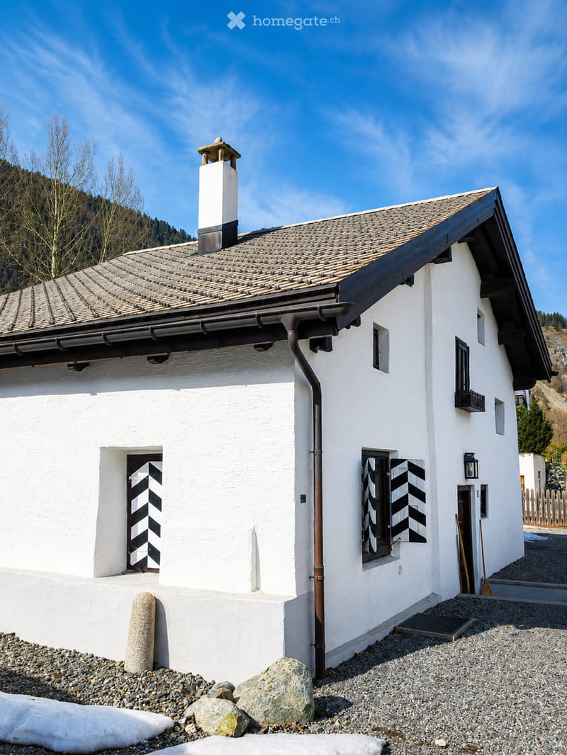 A white, single-story house with a tiled roof, chimney, and black-and-white striped shutters on the windows. The house is surrounded by gravel and rocks, with a wooden fence and trees in the background.