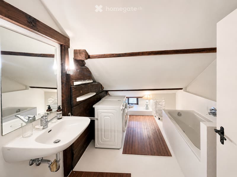 Bathroom with a sink, mirror, bathtub, washing machine, and a small lamp. Wooden beams and slanted ceiling are visible.