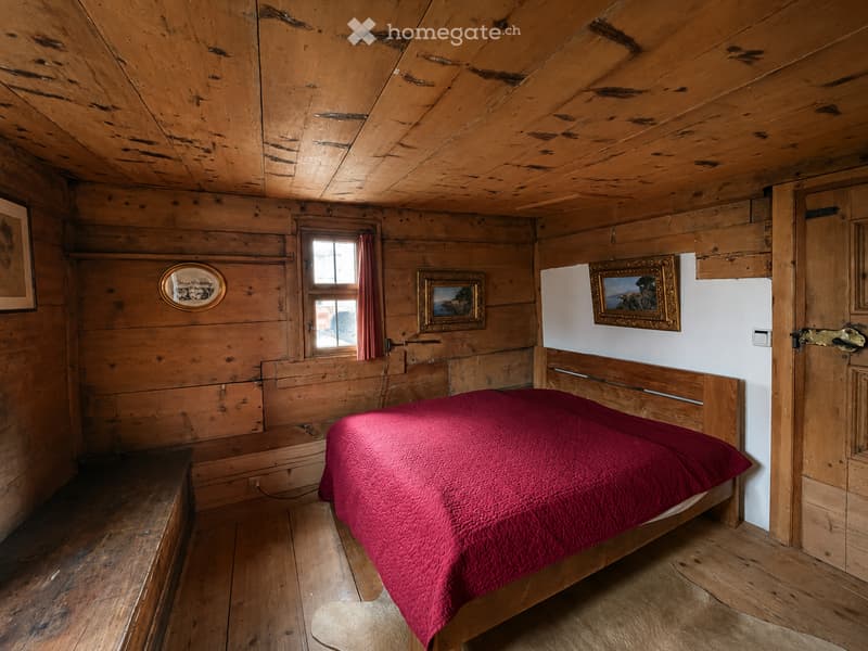 Cozy wooden bedroom with slanted wooden ceiling, red quilted bedspread, wooden bed frame, framed pictures on walls, and a window with curtains.