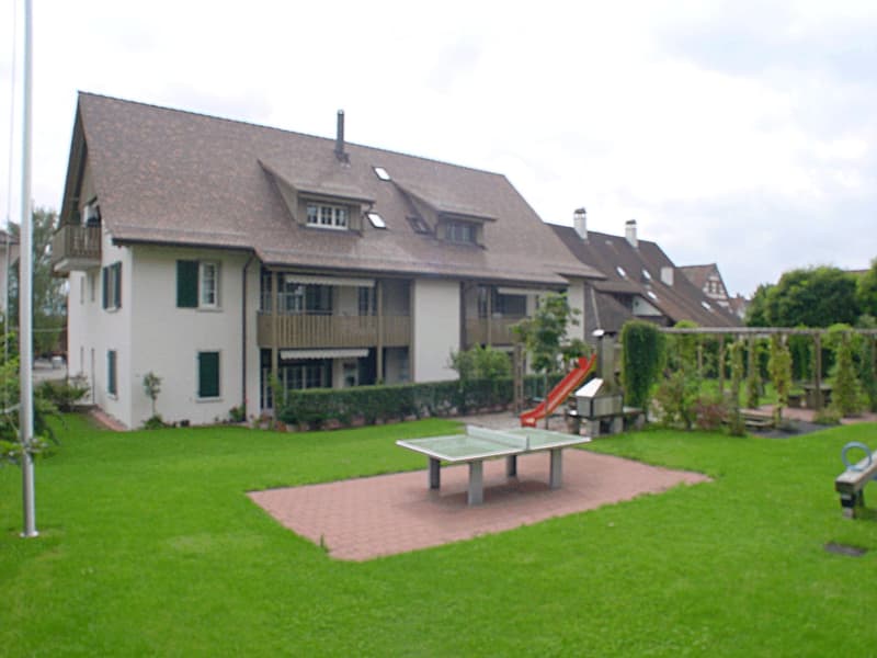 A well-maintained garden with a table tennis table, a red slide, and a bench, surrounded by a large house with multiple windows and balconies.