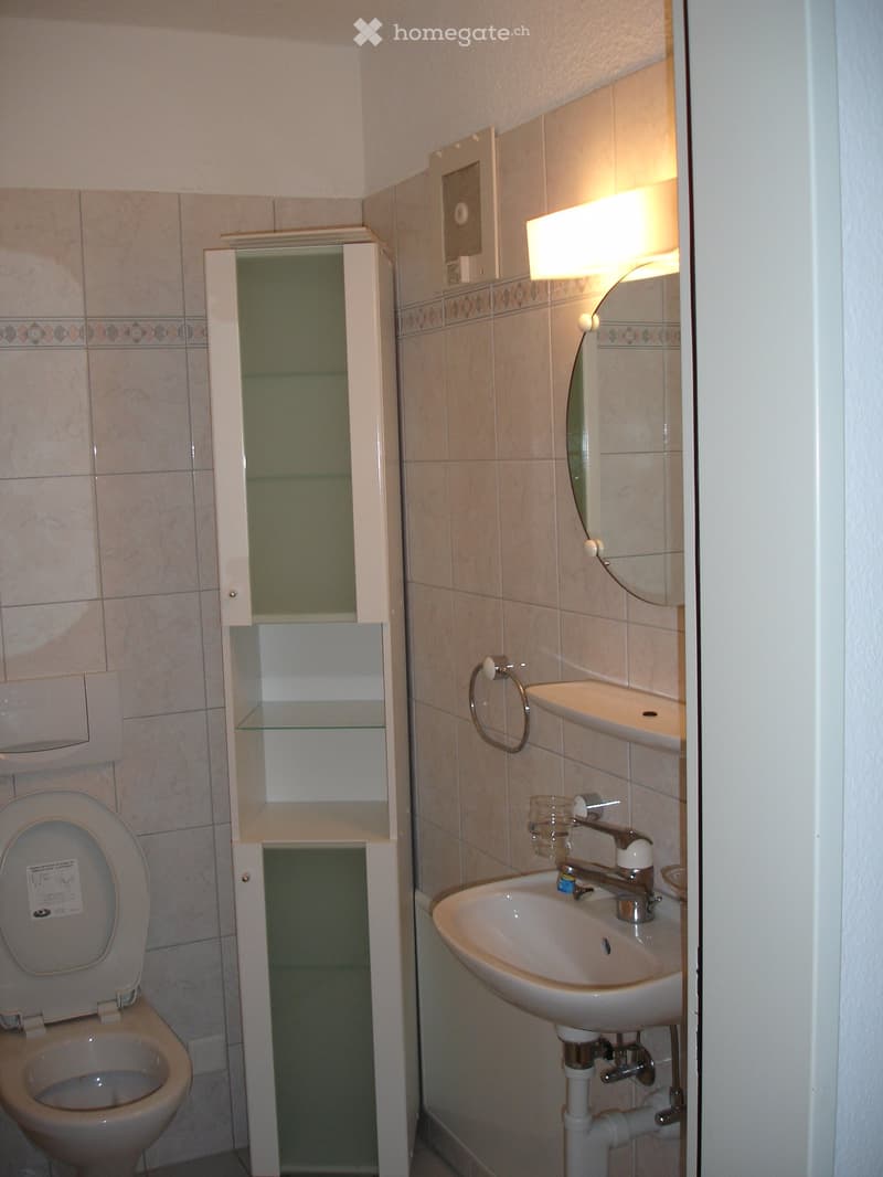 A small bathroom with a toilet, sink, and a mirror. The bathroom has white tiles on the walls and a white cabinet with glass doors.