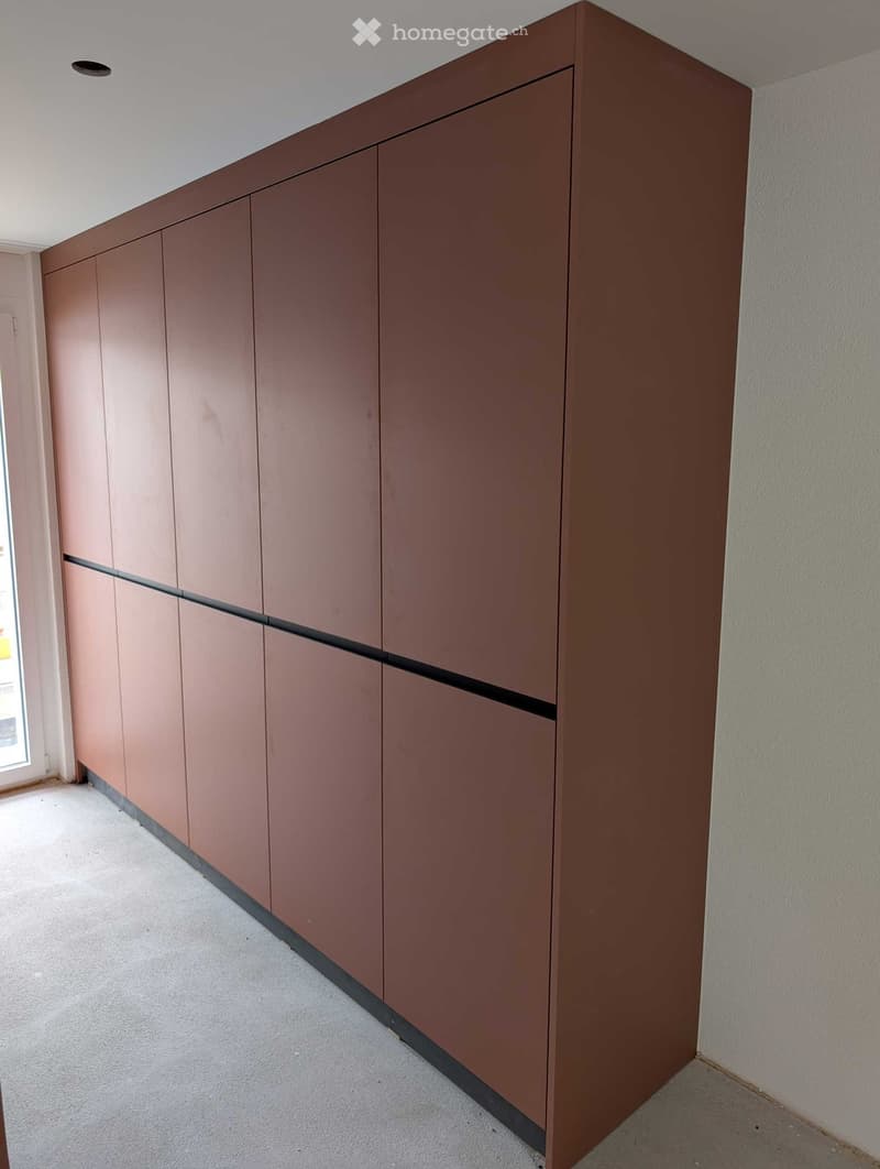 Empty room with brown built-in cabinets and a white wall. The floor is concrete, and there is a window on the left side.