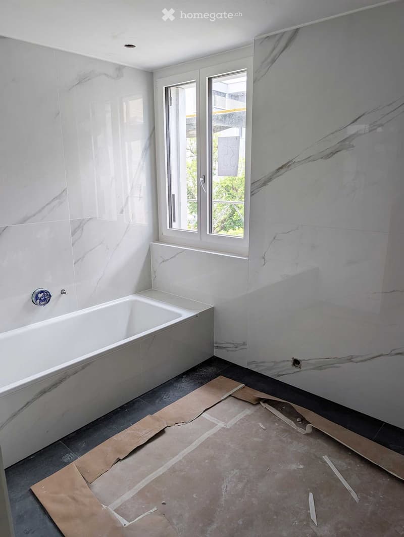 Empty bathroom with marble walls, a bathtub, and a window with frosted glass. The floor is unfinished with exposed concrete and cardboard sheets.