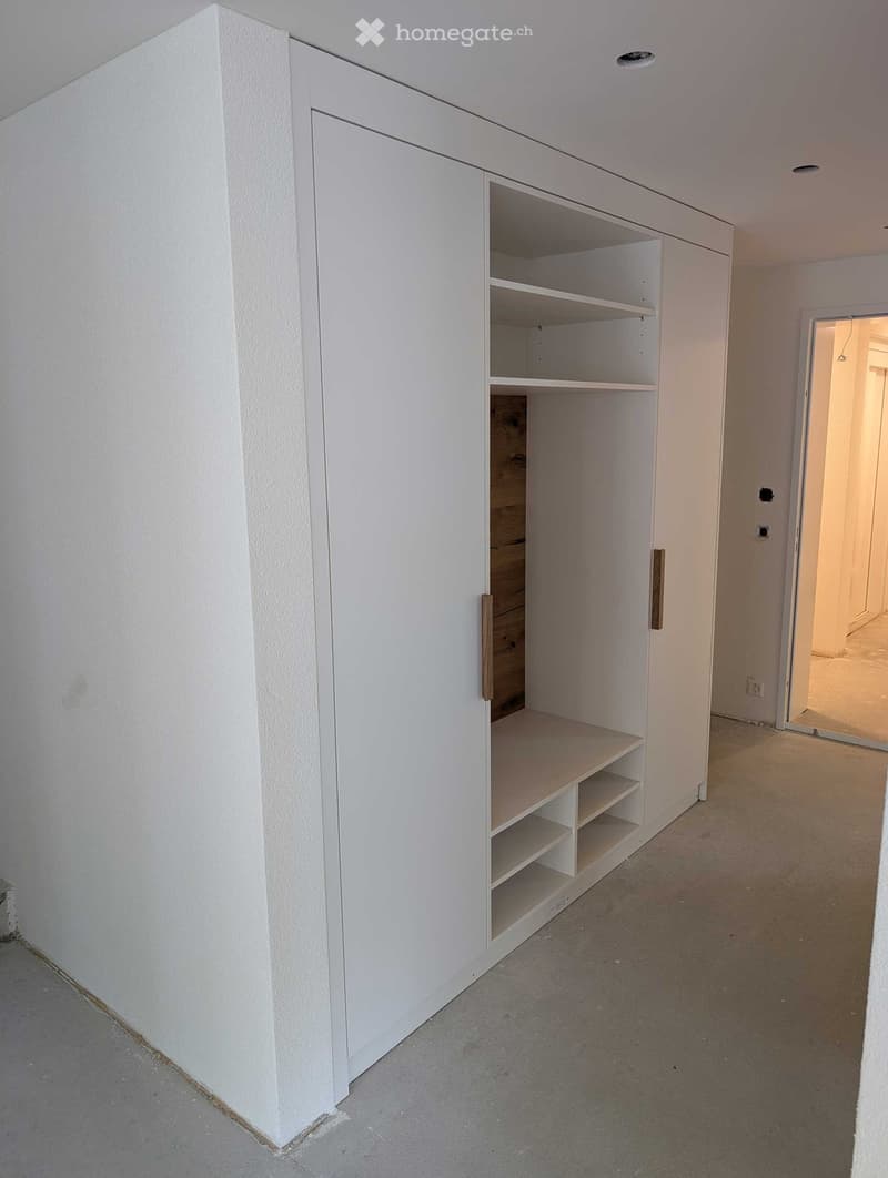 Empty room with white built-in shelving unit and open wooden drawer, concrete floor, white walls, and a doorway leading to another room.