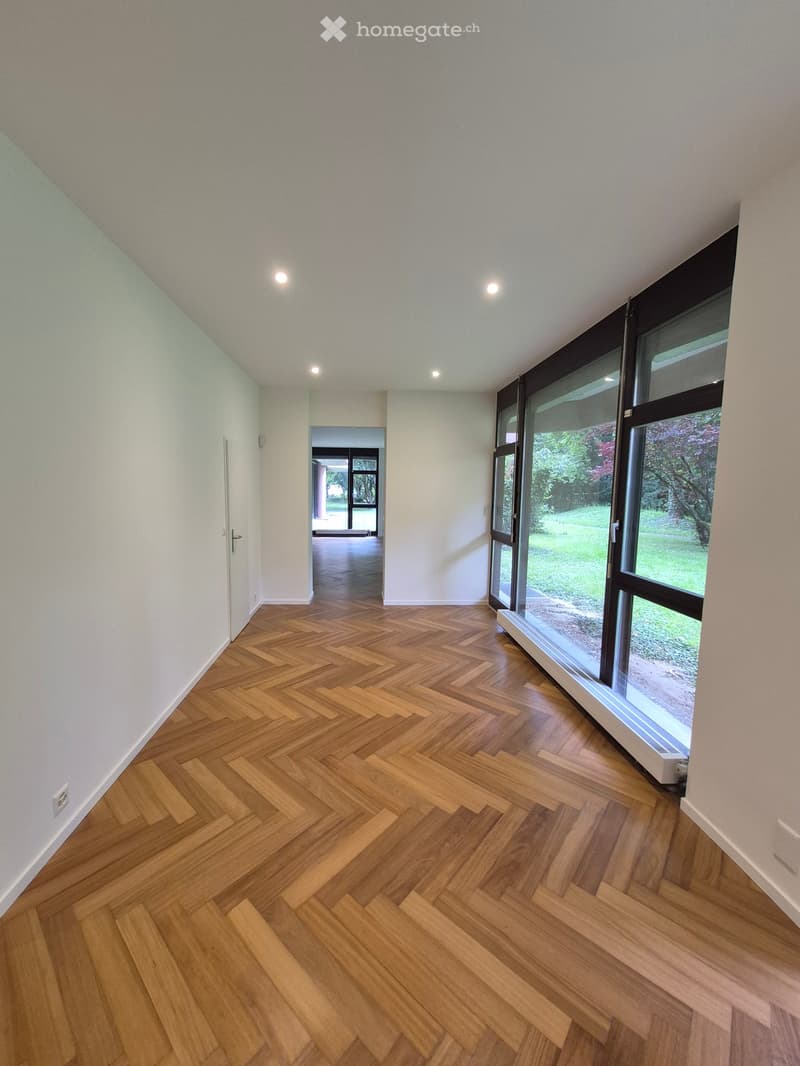 Empty hallway with herringbone wooden flooring, white walls, recessed lighting, and large windows overlooking a garden.
