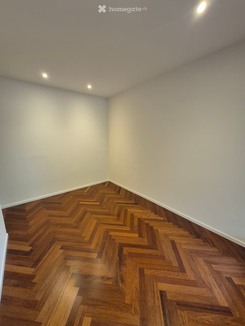 Empty room with light grey walls and a herringbone-patterned wooden floor. Recessed ceiling lights provide illumination.