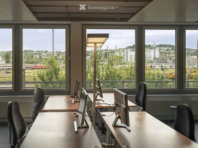 Office space with large windows, multiple desks, chairs, computers, and a floor lamp.