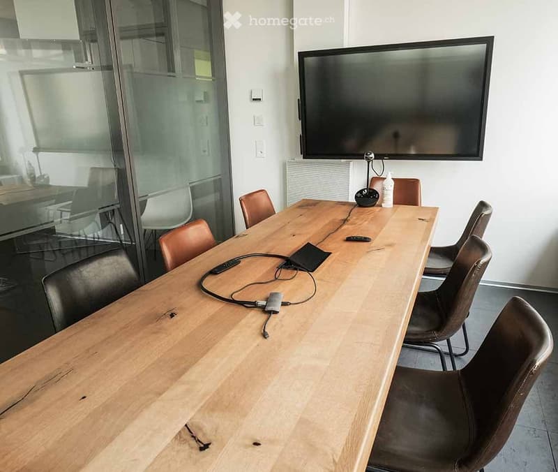 A modern meeting room with a long wooden table surrounded by brown leather chairs, a large flat-screen TV mounted on the wall, and a glass partition leading to another room.