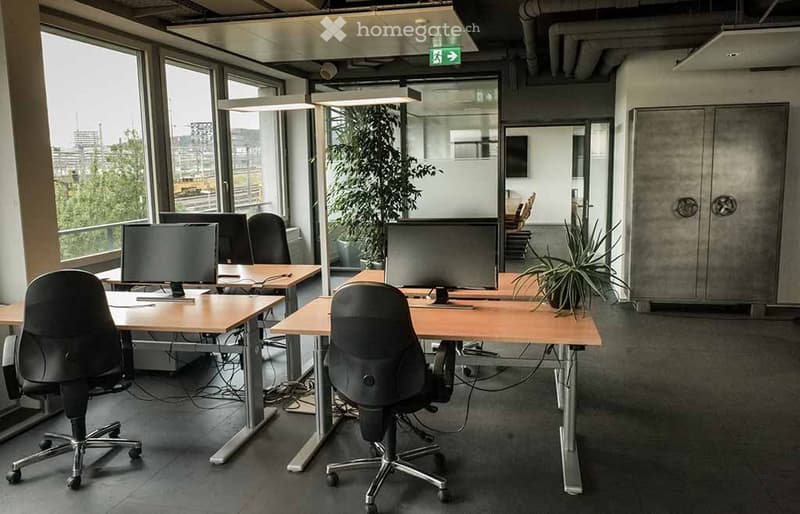 Modern office space with desks, chairs, computer monitors, and plants. Large windows provide natural light and a view of the city.