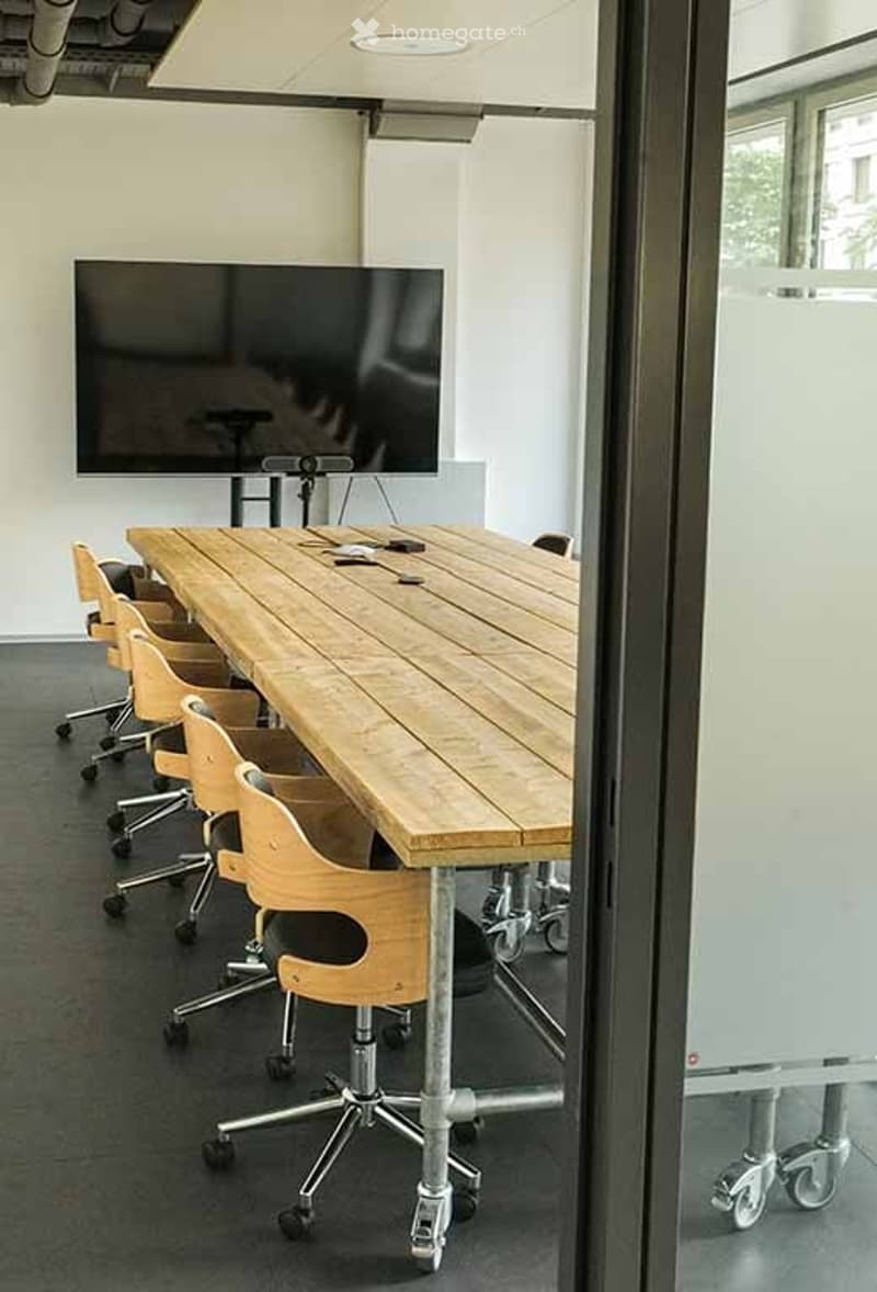 A modern conference room with a long wooden table surrounded by ergonomic chairs, a large flat-screen screen mounted on the wall, and a glass whiteboard on the side.