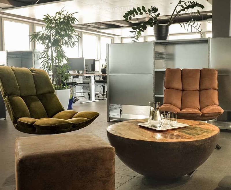 Modern office lounge area with a green leather sofa, brown leather chair, round wooden coffee table, glass cups, and a large potted plant.
