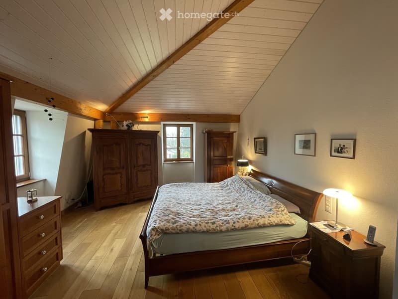Cozy attic bedroom with wooden beams, slanted ceiling, and wooden floor. Features a large bed with patterned bedding, nightstands with lamps, a wardrobe, and framed pictures on the wall.