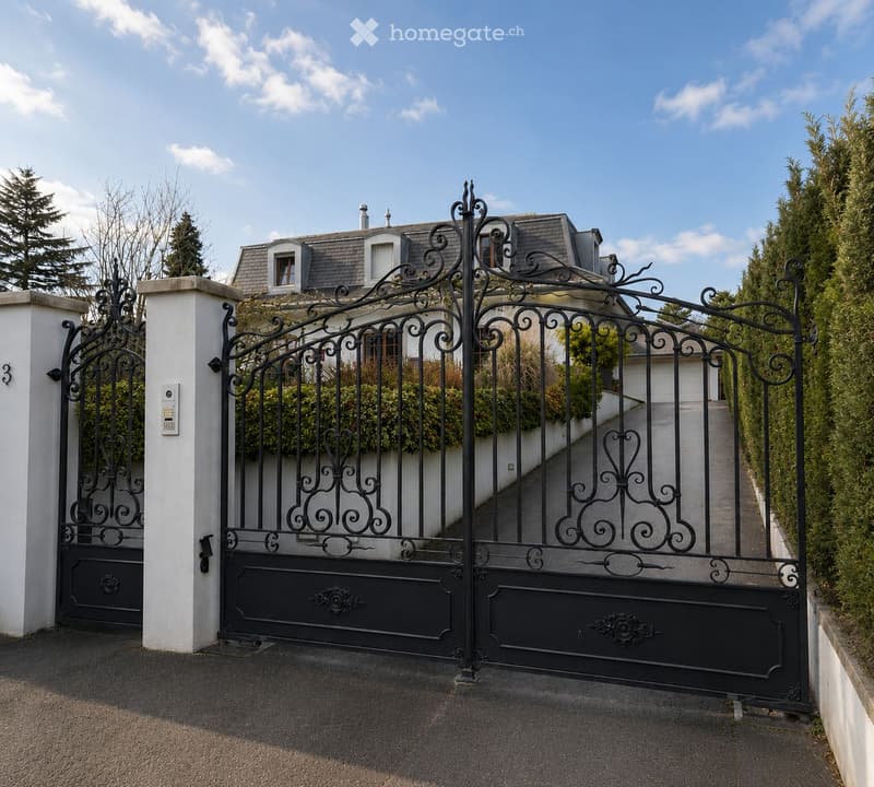 A large, elegant house with a grey roof and white walls is visible behind an ornate black wrought-iron gate. The gate features intricate scrollwork and is flanked by white pillars. The driveway leads up to the house, which has multiple windows and a small