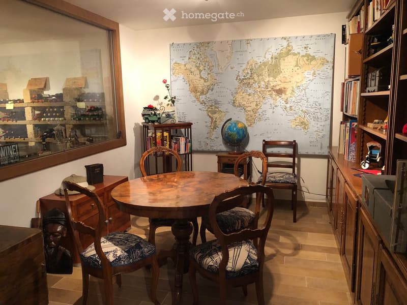 A cozy room with a round wooden table surrounded by four chairs, a bookshelf filled with books, a globe, a large world map, and a cabinet with a hat on top.