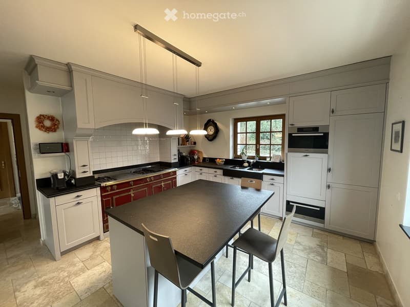 Modern kitchen with white cabinets, a central island, and stainless steel appliances. Features include a gas stove, refrigerator, microwave, and a sink with a window above. The island has two bar stools and is surrounded by light-colored tiles.