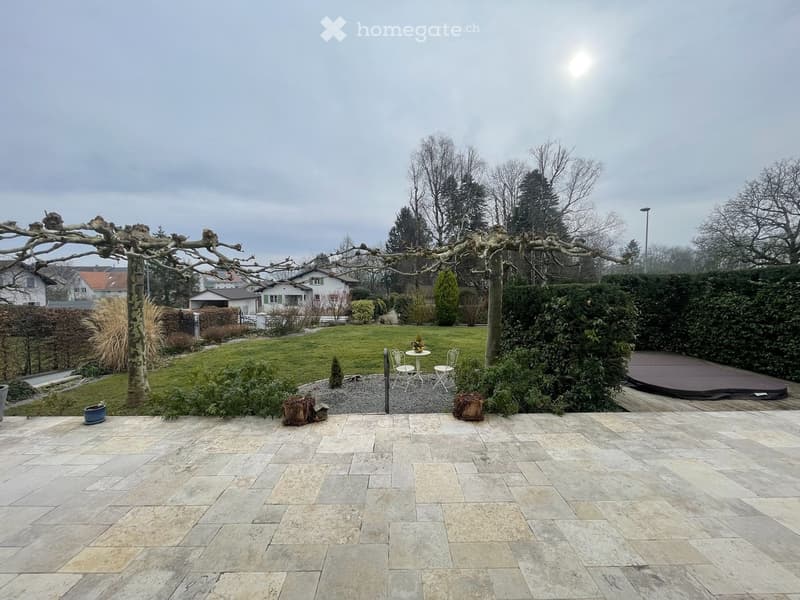Garden with a stone patio, small seating area with a table and chairs, surrounded by greenery and trees.