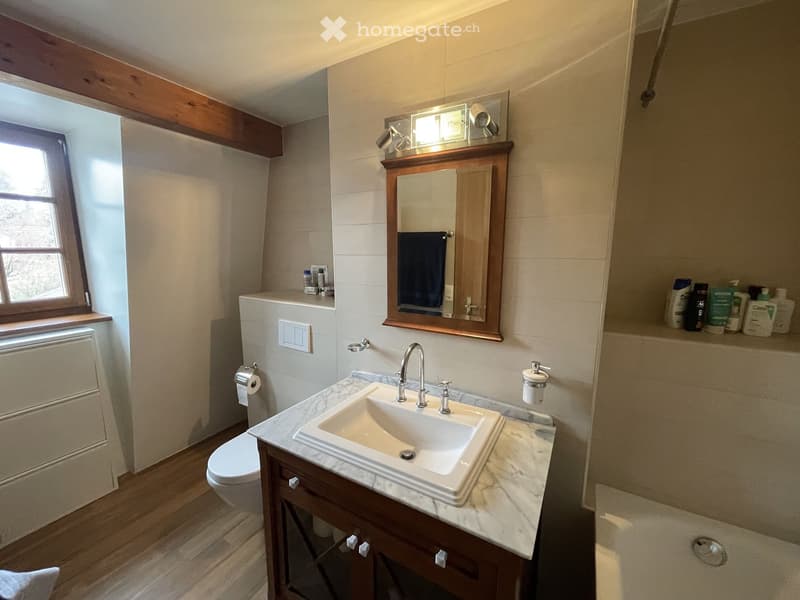 Modern bathroom with a white marble countertop, a single sink, a mirror with wooden frame, a toilet, and a shower area. The room features wooden flooring and a window letting in natural light.