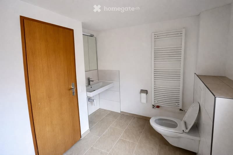 Modern bathroom with a toilet, sink, mirror, radiator, and cabinet. The space features tiled flooring and white walls.