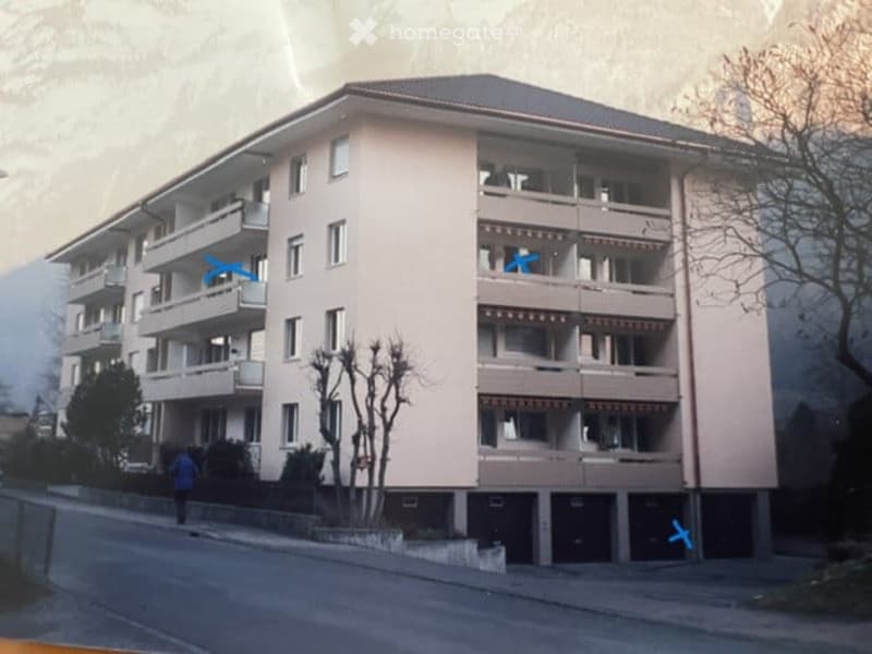 Multi-story residential building with multiple balconies and windows, surrounded by trees and a road.