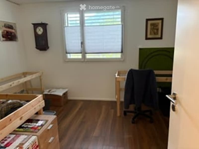 A small, furnished room with wooden flooring, a desk, a chair, a bed, and a window with blinds. There is a clock on the wall and some books on the floor.